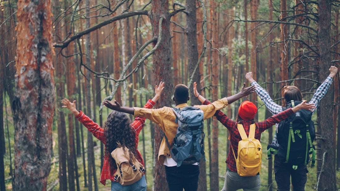 Four friends with backpacks raise arms in forest.